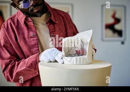 Uomo che indossa occhiali da sole e camicia rossa che guarda da vicino una scultura contemporanea in una galleria d'arte moderna con opere d'arte incorniciate sullo sfondo Foto Stock