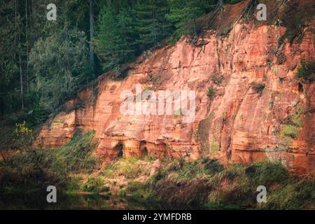 Scogliera di arenaria rossa vivace adornata con muschio e vegetazione, circondata da alberi e riflessa in un tranquillo fiume sottostante. Foto Stock