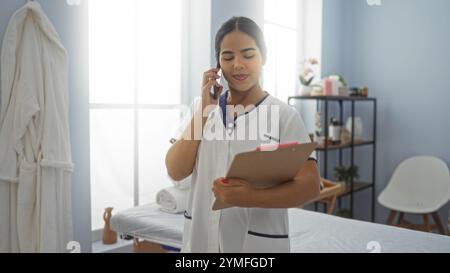 Donna nel centro benessere che indossa un'uniforme al telefono e tiene gli appunti in una tranquilla sala benessere. Foto Stock