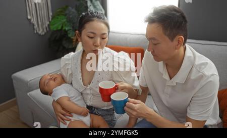 Madre e padre seduti insieme in un accogliente soggiorno tenendo tazze da caffè mentre il loro bambino dorme pacificamente tra le sue braccia Foto Stock