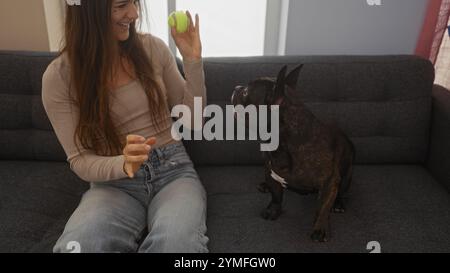 Una giovane donna ispanica che gioca con il suo bulldog francese in un accogliente soggiorno di un appartamento. Foto Stock