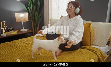 Una donna anziana siede su un letto in un'accogliente camera da letto, impegnata in meditazione, mentre un animale domestico chihuahua nelle vicinanze cattura l'atmosfera tranquilla al coperto. Foto Stock