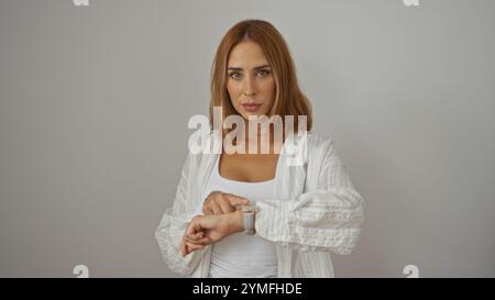Giovane donna che guarda il suo smartwatch su una parete isolata di sfondo bianco, mostrando le sue belle e attraenti caratteristiche come un brun ispanico adulto Foto Stock