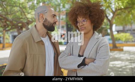 Una coppia interrazziale si trova in un parco cittadino, apparendo impegnata in una conversazione seria, circondata da vegetazione urbana ed elementi di strada. Foto Stock