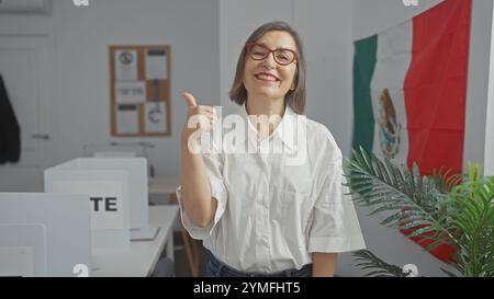 Una donna di mezza età regala un pollice in una sala di voto messicana con una bandiera sullo sfondo. Foto Stock