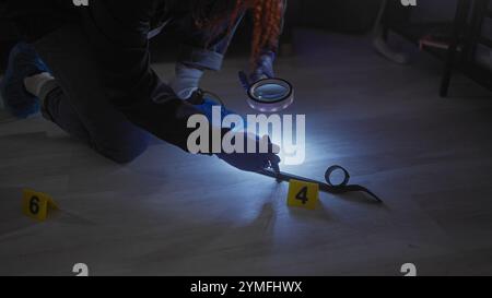 Una donna esamina le prove su una scena del crimine al chiuso poco illuminata usando una lente d'ingrandimento e indossando guanti. Foto Stock