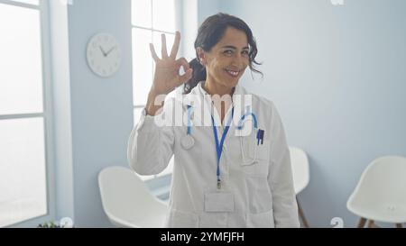 Una dottoressa ispanica sicura di sé dà un segnale di OK in una stanza clinica luminosa, segnalando il successo o la soddisfazione. Foto Stock