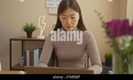 Una giovane donna cinese lavora sul suo computer portatile in un accogliente soggiorno decorato con piante e libri. Foto Stock