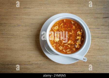 Zuppa cinese calda e acida con brodo di pollo con uova, carne e verdure, ciotola bianca con cucchiaio di porcellana su un tavolo di legno in un ristorante asiatico, Foto Stock