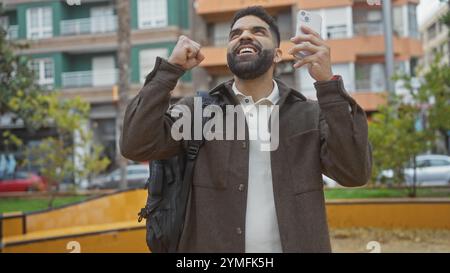 Un allegro ispanico festeggia mentre tiene in mano uno smartphone in un parco urbano, trasudando uno stile di vita moderno e informale. Foto Stock