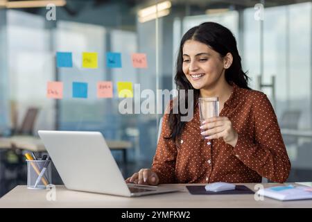 Una donna d'affari latinoamericana tiene un bicchiere d'acqua, sorridendo al laptop in ufficio. L'immagine evidenzia idratazione, concentrazione e positività nell'ambiente di lavoro con un ambiente moderno e collaborativo. Foto Stock