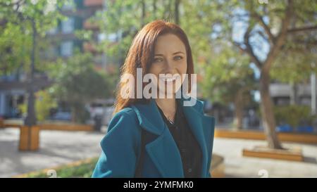 Donna rossa sorridente che indossa un cappotto blu in un parco urbano soleggiato Foto Stock