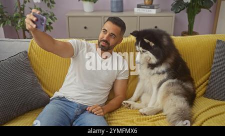 Un bel giovane ispanico si fa un selfie con il suo cane husky su un divano giallo in casa. Foto Stock