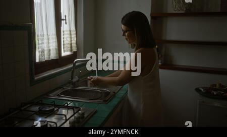 Una giovane donna ispanica si lava le mani ad un lavandino in una vecchia casa italiana con un tipico ambiente da cucina. Foto Stock