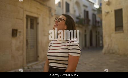 Una bella giovane donna ispanica sorride in un'affascinante stradina di lecce, puglia, italia, circondata da un'architettura storica su una soleggiata strada Foto Stock