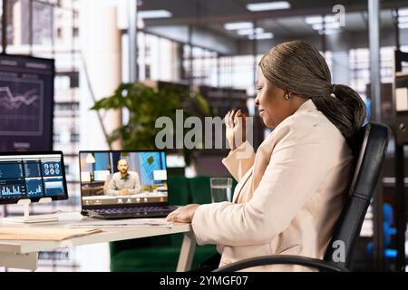 Team leader in un ufficio moderno che parla con il manager regionale in una riunione di teleconferenza. Donna d'affari afroamericana alla scrivania del luogo di lavoro in videocall online con dirigente Foto Stock