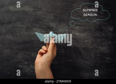 Una mano che tiene una barca di carta su una lavagna con sopra le parole Summer Season. Concetto di divertimento e svago, dato che la barca di carta è un simbolo Foto Stock