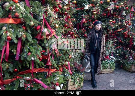 Londra, Regno Unito - 19 novembre 2024. Una giovane donna con un cappotto di pelliccia sintetica con stampa leopardata si posa davanti agli alberi di Natale decorati al Covent Garden, una popolare Foto Stock