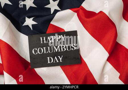 Un cartello nero con la scritta "Happy Columbus Day" è posto sopra una bandiera americana bianca e rossa Foto Stock