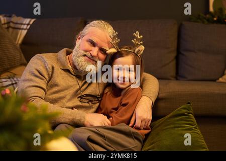 Nonno con barba bianca e capelli grigi che abbracciano la nipote che indossa corna di renna mentre si siede sul divano in un accogliente soggiorno durante le vacanze Foto Stock