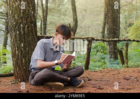 Ritratto di un uomo asiatico sicuro che scrive su un taccuino mentre pensa e guarda lontano nella foresta naturale. Benessere sanitario tranquillo e rilassante Foto Stock