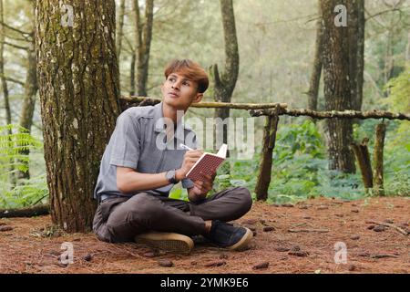 Ritratto di un uomo asiatico sicuro che scrive su un taccuino mentre pensa e guarda lontano nella foresta naturale. Benessere sanitario tranquillo e rilassante Foto Stock