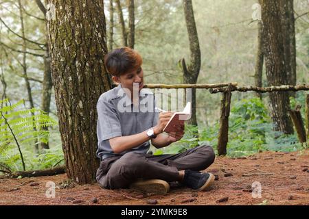 Ritratto di un uomo asiatico sicuro che scrive su un taccuino mentre pensa e guarda lontano nella foresta naturale. Benessere sanitario tranquillo e rilassante Foto Stock