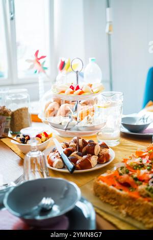 Una colazione ben organizzata con un piatto di frutta, pasticcini e pretzel su un tavolo da pranzo illuminato dal sole. Perfetto per riunioni o per un piacevole soggiorno Foto Stock