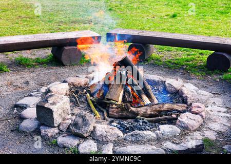 Fuoco acceso nel caminetto sullo sfondo di panche di legno e erba verde. Area grigliata. Foto Stock