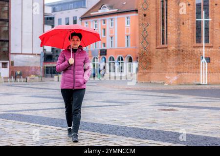 Una donna di mezza età sotto un ombrello rosso attraversa la piazza. Le gocce di pioggia sono visibili. Foto Stock