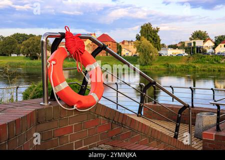 Un salvagente arancione è appeso alla ringhiera di un molo sulla riva del fiume. Foto Stock