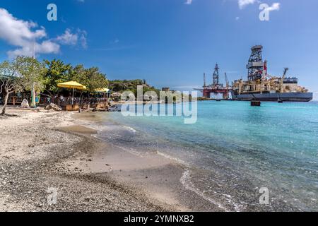 Il ristorante sulla spiaggia Brisa do Mar si trova a Caracasbaaiweg, Jan Thiel, Curacao, con acque turchesi dei Caraibi e una spiaggia di sabbia bianca. Sullo sfondo a destra, un grande impianto di perforazione offshore è ancorato a Caracasbaai, un contrasto tipico della costa orientale di Curacao tra l’idilliaca spiaggia turistica e l’industria petrolifera e del gas. Ristorante Brisa do Mar. Kòrsou Foto Stock
