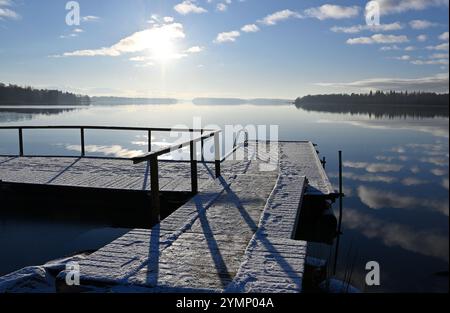 una tranquilla vista sul lago nelle limpide mattine d'inverno. Un molo di legno spolverato di neve si estende nell'acqua calma e riflettente, creando un ambiente tranquillo e sereno Foto Stock