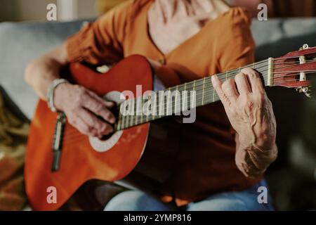 Persona anziana che suona la chitarra acustica seduto comodamente. Vista ravvicinata che evidenzia i movimenti delle mani e la concentrazione sullo strumento musicale Foto Stock