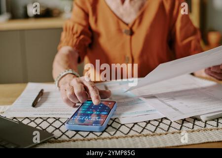 Donna anziana che lavora con documenti e smartphone, gestisce i registri finanziari a casa. Senior che si concentra sulla gestione di bollette e rendiconti finanziari sul tavolo Foto Stock
