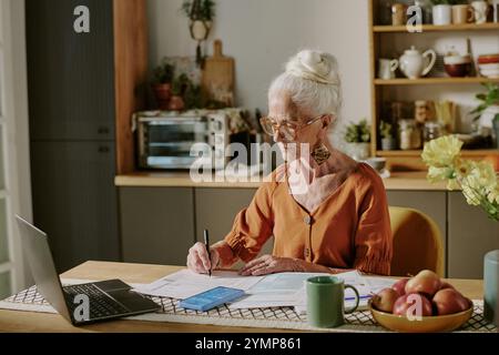 Donna anziana che indossa gli occhiali in un'accogliente cucina che lavora su documenti con laptop e smartphone sul tavolo, circondata da un arredamento accogliente con fiori e frutta Foto Stock
