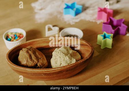 Ciotola in legno con impasto per biscotti e varie forme per taglierine e caramelle colorate sul tavolo, creando la configurazione ideale per le attività di cottura fatte in casa Foto Stock