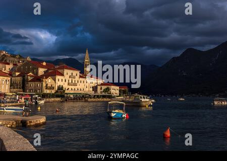 La vista оf di Perast, Montenegro, è illuminata dalla calda luce dorata del sole che tramonta. Il primo piano è sereno, con barche che galleggiano, mentre il buio st Foto Stock