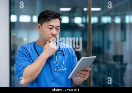 Uomo asiatico medico che indossa scrub blu utilizza tablet digitale nel moderno ufficio medico. Espressione focalizzata indica considerazione e analisi. Concetto di tecnologia nel settore sanitario. Foto Stock