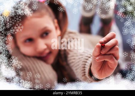 Una ragazza sorridente giace sul pavimento, mostra una decorazione natalizia rossa, circondata da fiocchi di neve che cadono, catturando la gioia e la magia del mare natalizio Foto Stock