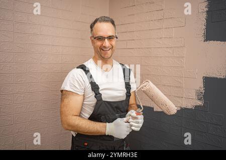 Un uomo che indossa una camicia bianca e una tuta nera è in piedi con un rullo per pittura in mano. Sembra essere pronto a dipingere una superficie, con i suoi strumenti Foto Stock