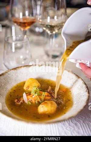 Il cameriere versa il brodo in una ciotola di zuppa con gnocchi, carote ed erbe aromatiche, in un raffinato ristorante Foto Stock