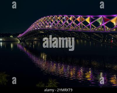 Moderno ponte pedonale illuminato da vivaci luci viola che si riflettono sulle acque calme di notte Foto Stock