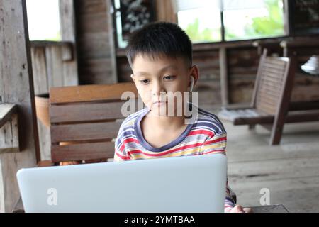 Un ragazzo siede in un interno rustico, concentrato sul laptop mentre indossa le cuffie. L'ambiente accogliente mette in risalto il suo impegno con la tecnologia e. Foto Stock