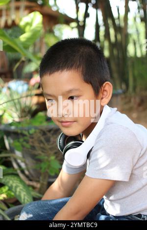 Un bambino siede all'aperto circondato da una vegetazione lussureggiante e indossa le cuffie. La sua premurosa espressione riflette un momento di pace e di piacere in natura Foto Stock