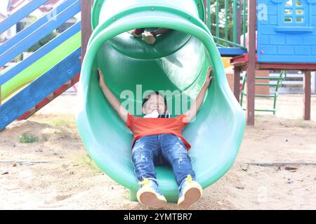 Un bambino allegro scivola giù su uno scivolo verde del parco giochi, incarnando lo spirito di divertimento e spensieratezza. I colori luminosi e l'ambiente esterno esaltano la figura Foto Stock