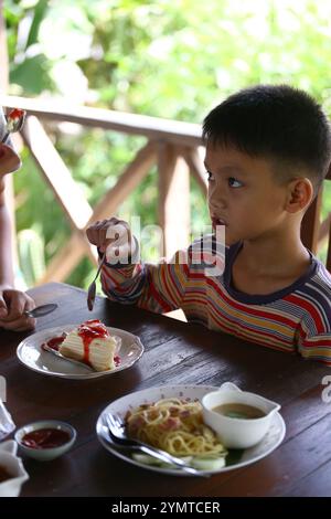 Un ragazzo si diverte con un delizioso dessert seduto a un tavolo di legno. La sua premurosa espressione mette in risalto la gioia di condividere un pasto in un piacevole Foto Stock