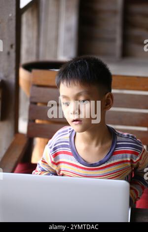 Un ragazzo siede comodamente su una sedia di legno, profondamente concentrato sul suo computer portatile. L'ambiente intimo della casa mette in risalto la sua curiosità e il suo coinvolgimento Foto Stock