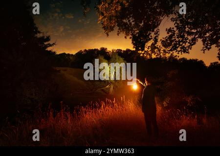 Ripresa notturna di un uomo in piedi in un paesaggio rurale, che tiene la luce e contempla la natura. Con erba e alberi illuminati Foto Stock