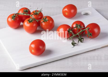 I pomodori ciliegini sono esposti su un tagliere bianco con alcuni sulla superficie e altri ancora attaccati alla vite, evidenziandone la freschezza e la freschezza Foto Stock